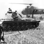 A helicopter and tank in the background with soldiers on top of it.