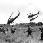 A group of soldiers in the field with helicopters flying overhead.